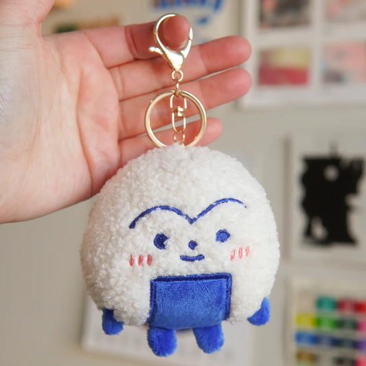Plush keychain with an onigiri cartoon character design held by a hand against a blurred background.
