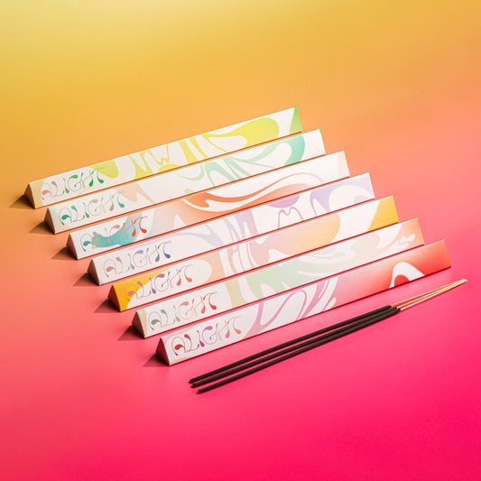 Six triangular prism boxes with colorful abstract swirl patterns are arranged diagonally on a yellow-to-pink gradient background, with three black incense sticks placed in front.