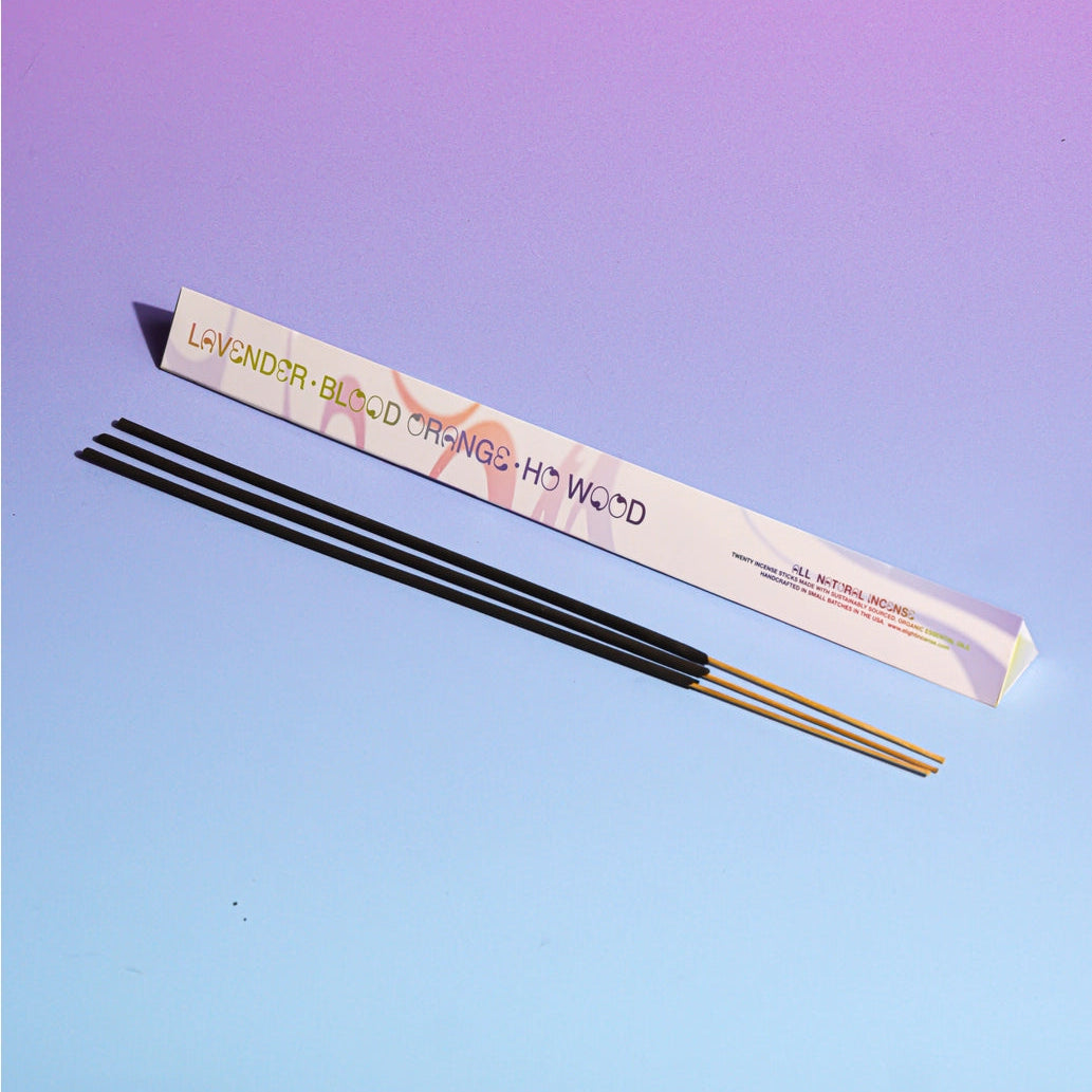 Three incense sticks in front of a long white box with colorful swirls labeled 'LAVENDER • BLOOD ORANGE • HO WOOD' on a lavender-to-blue gradient background.