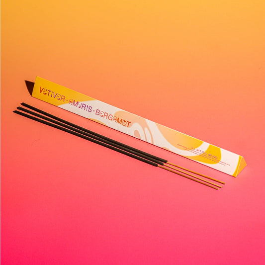 Three incense sticks in front of a long,  white and yellow box labeled 'Vetiver • Amyris• Bergamot' on a vibrant yellow-to-pink gradient background.