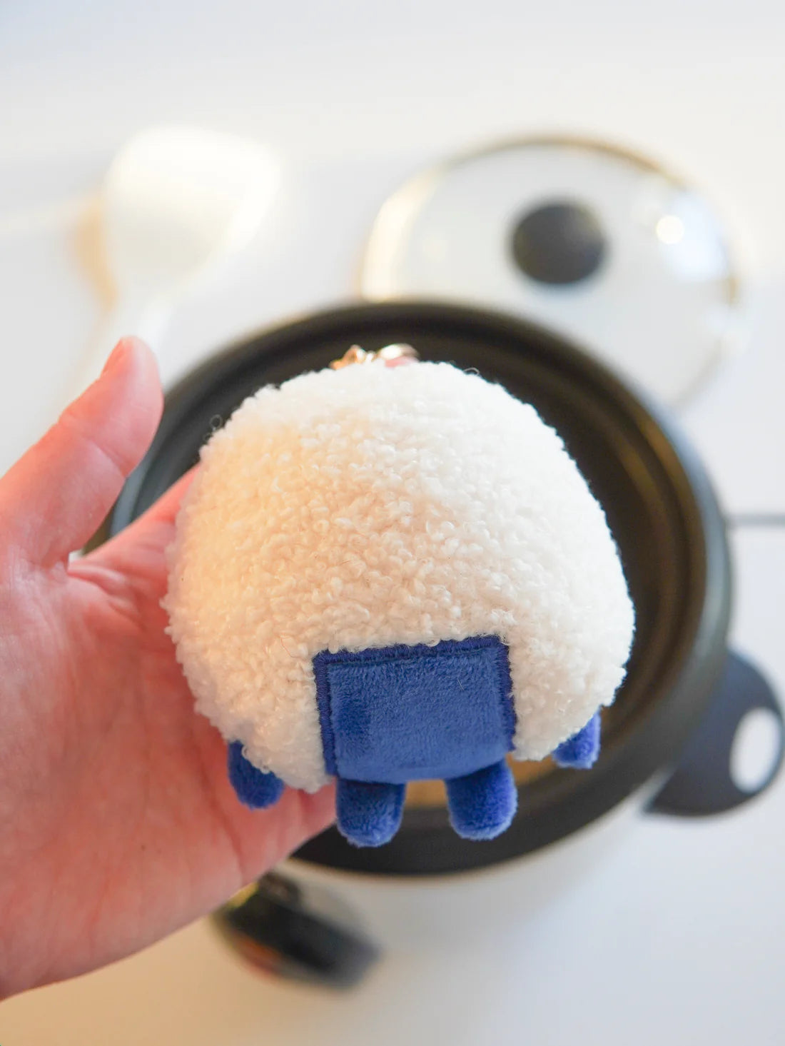 Plush toy resembling a rice ball with a blue patch as the "seaweed" held over a rice cooker.