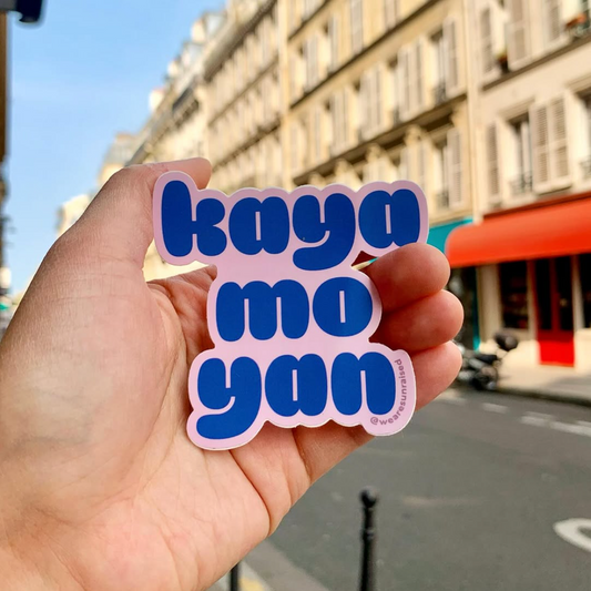 Hand holding a pink and blue sticker with 'kaya mo yan' text against a city street background